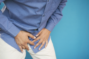 A man wearing a blue, button up shirt and khaki pants, is bent over in pain. He is clutching is groin.
