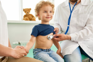 A small child getting a medical check-up. He is sitting on a doctor's table, in-between two adults. One adult is holding the boy's shirt up and listening to his chest with a stethoscope. The other adult is holding the child's hand.