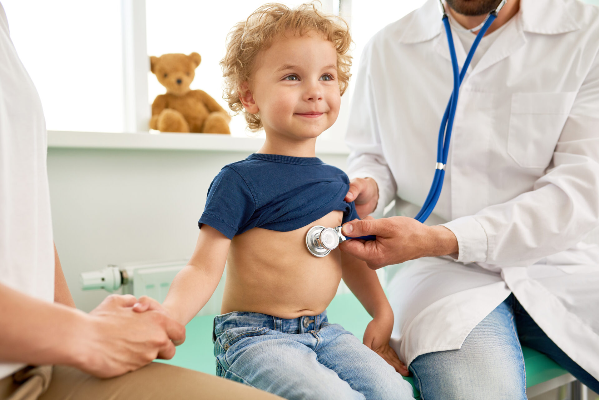 A small child getting a medical check-up. He is sitting on a doctor's table, in-between two adults. One adult is holding the boy's shirt up and listening to his chest with a stethoscope. The other adult is holding the child's hand.