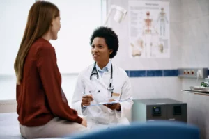 A brunitte woman sitting on an examination table in a doctor's office, ready for a well-woman exam. An African American woman sits on a stool next to the table.