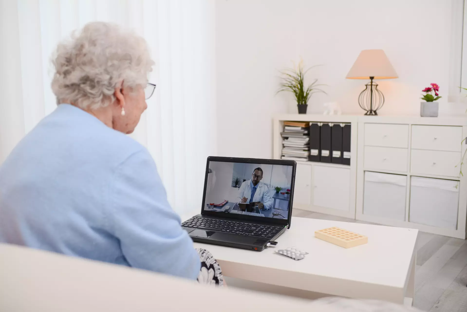 An elderly woman sits on a white couch in a living rom, using her laptop to speak to a doctor using Telemedicine.