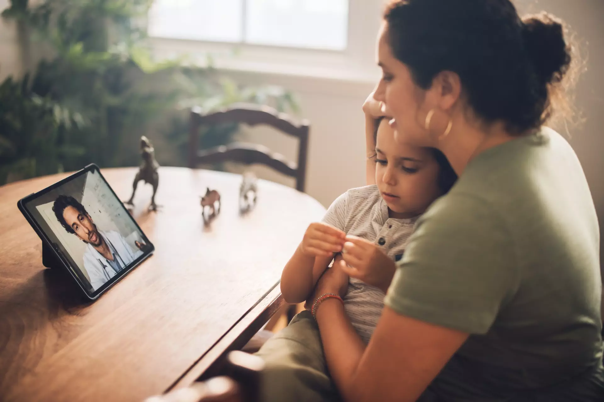 A woman holds a small child in her lap while she uses Telemedicine to communicate with a doctor on a tablet set on a dining room table, next to some toy dinosaurs.